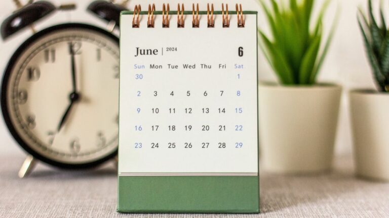 Desk calendar that reads June 2024 sits centered on desk with plants and a retro bell alarm clock blurred in background