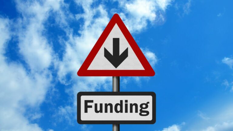 street style road sign on metal pole against a blue sky with wispy clouds. On top of pole is white triangle with redoutline and black arrow pointing down in centre. Below that is a white rectangle with black outline that reads funding in black