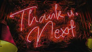 red neon sign reads: Thank u, Next against a dark background of fake plants and bordered by dimly lit retro-ish homestyle lamps