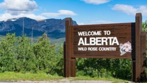 Large brown wooden sign on side of rural road reads welcome to Alberta wild rose county in white, with decorative reose icon. Background features green landscape, mountains and blue sky with wispy clouds