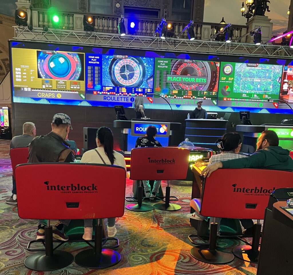 Personas jugando en una sala de casino equipada con pantallas gigantes que muestran mesas de ruleta, craps y blackjack.