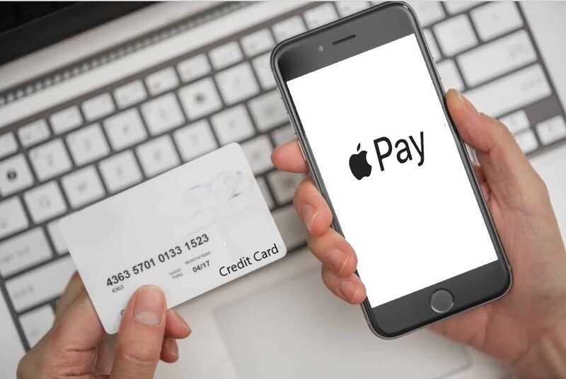 Manos sosteniendo una tarjeta de crédito blanca y un teléfono inteligente que muestra el logo de Apple Pay, sobre un teclado de computadora blanco en el fondo.