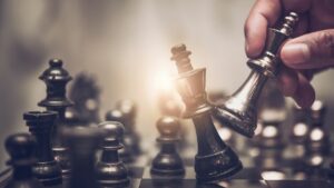 Backlit close up of chess board. Hand holding royal chess piece knocks over competitor piece