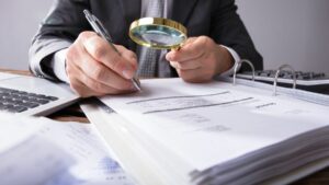Close up of a desk covered in receipts and files. A masculine coded business person examines a binder of receipts with a magnifying glass