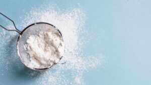 close up of a metal strainer holding flour that is dusted over a pale blue background surface