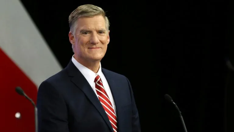 former state senator Andy Sanborn stands on dark stage looking towards camera wearing dark blue suit with white shirt and red and white diagonal striped tie