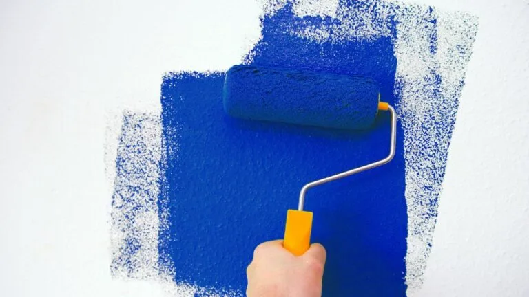 Hand holding orange handled paint roller rolls royal blue paint on white wall