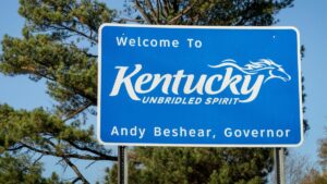Blue and white highway road sign redas Welcome to Kentucky, unbridled spirit, Andy Beshear, Governor. Blue sky and trees fill background
