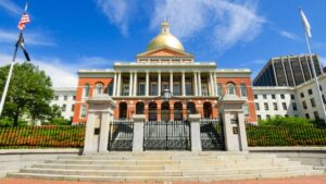 Massachusetts State House on a sunny day with blue skies and sparse, wispy clouds. Photo is framed by flag poles.