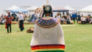 Native American in full tribal regalia stands with back to camera at Chumash Intertribal Powow in Malibu, CA in 2024. Blurred Powwow grounds fill background