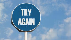 Round blue and while street sign on white pole reads try again against blue sky with wispy white clouds