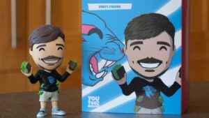 MrBeast You Toox vinyl figure golding fast stacks of cash sits on table next to retail box with figure's image. Wood paneled walls fill background