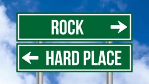 Pair of horizontal green and white road signs. Top reads rocks with right arrow, bottom reads hard place with left arrow. Blue cloudy sky fills background