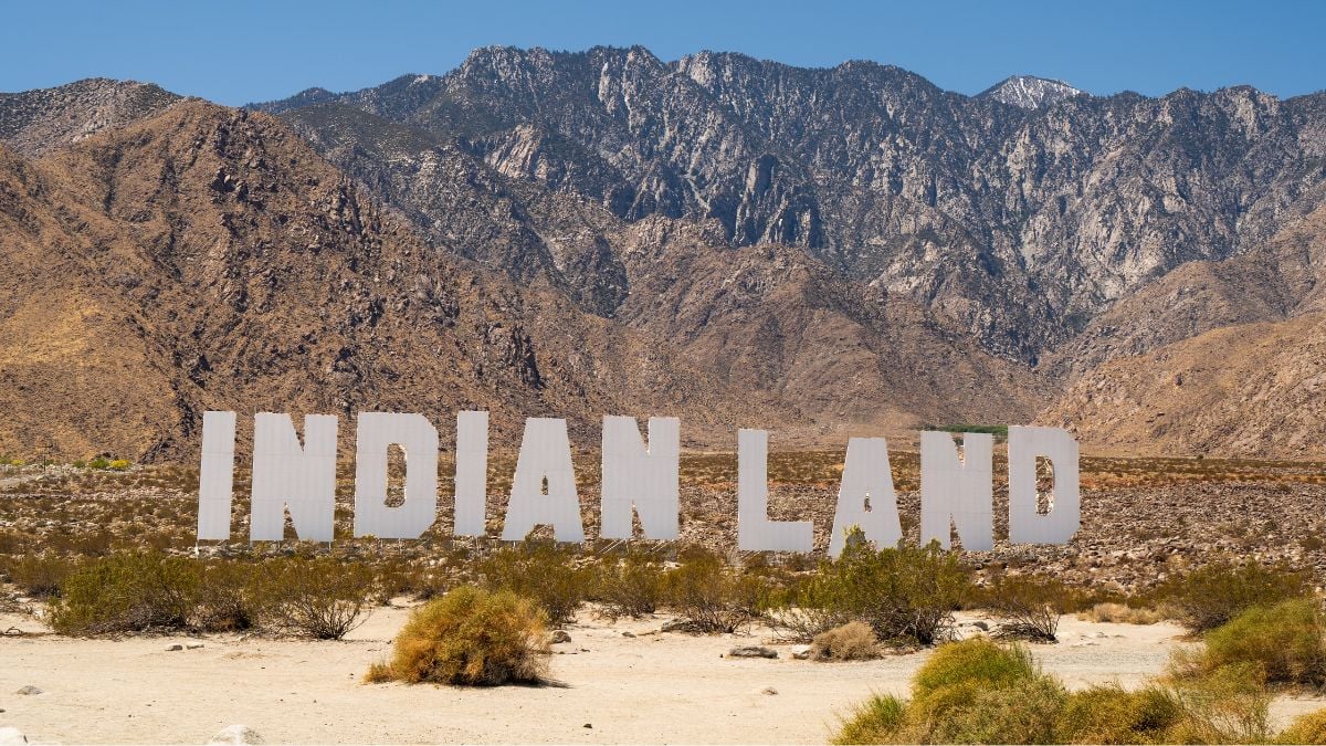 Desert land in Palm Springs, CA fearures hollywood-style sign that reads Indian Land with mountains and blue sky filling the background