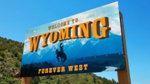 Painted roas/state sign reads 'Welcome to Wyoming, Forever West.' Sign features shadow of a cowboy on a bucking horse with mountains in the backdrop. Background behind sign is filled with a solid blue sky and rolling hills