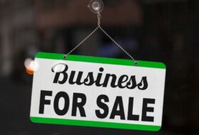 Black and white sign hanging from suction cup on glass door. Top and bottom margins of sign are green. Sign reads business for sale