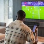 man holding smartphone with sports bets app while watching football match