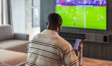 man holding smartphone with sports bets app while watching football match