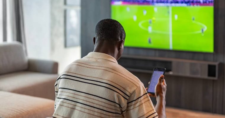 man holding smartphone with sports bets app while watching football match