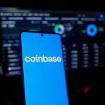 Coinbase logo on a smartphone in front of a trading screen setup