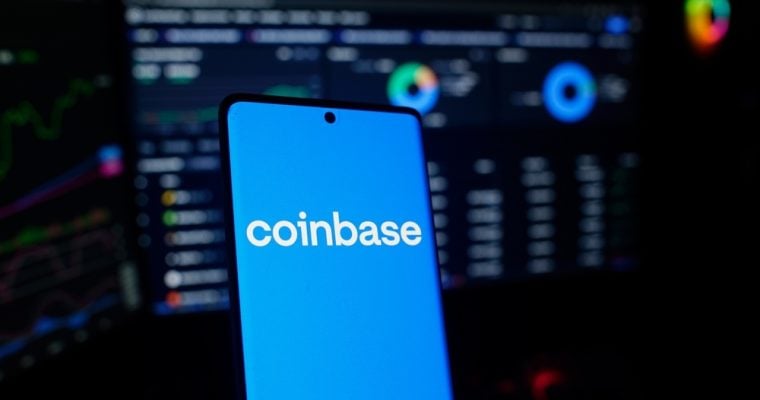 Coinbase logo on a smartphone in front of a trading screen setup