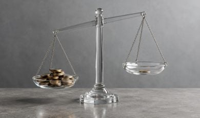 A clear glass scale sits on a gray surface with a heavy pile of gold coins on one side and a single coin on the other.