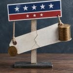 A conceptual image shows a cracked blank wooden sign hanging like a scale from a red, white, and blue patriotic banner, balanced by a stack of coins.