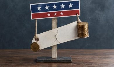A conceptual image shows a cracked blank wooden sign hanging like a scale from a red, white, and blue patriotic banner, balanced by a stack of coins.