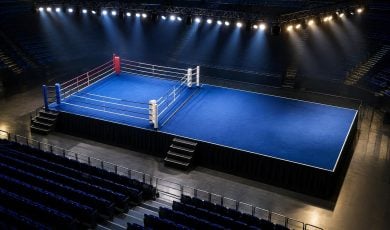 An empty boxing ring with a blue canvas stands in the center of a dark, tiered arena illuminated by overhead spotlights.