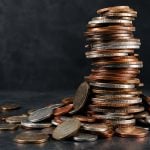A large stack of mixed copper and silver colored coins sits on a dark surface, surrounded by loose coins.