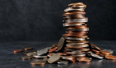 A large stack of mixed copper and silver colored coins sits on a dark surface, surrounded by loose coins.