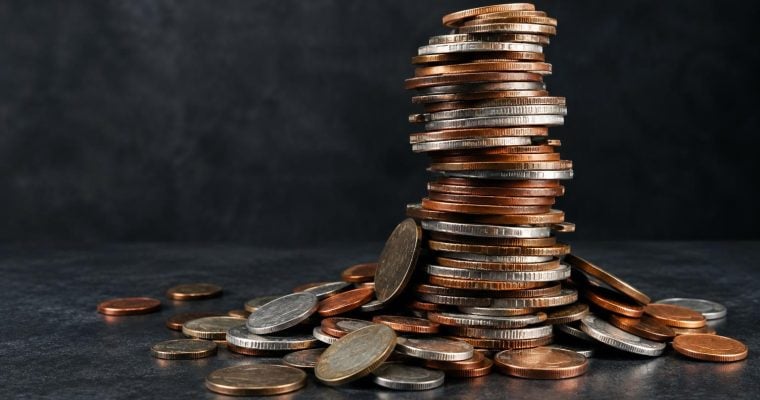 A large stack of mixed copper and silver colored coins sits on a dark surface, surrounded by loose coins.