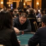 A group of people sitting around a green felt table playing a game of poker in a crowded, dimly lit casino hall.
